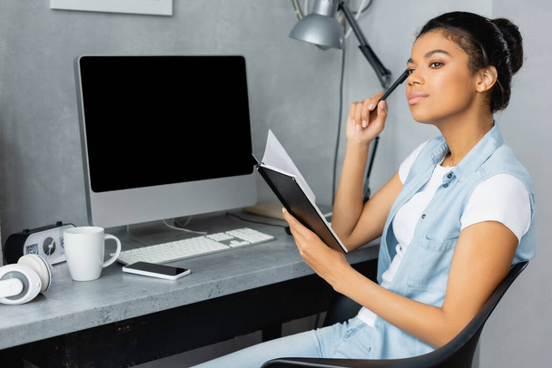 pensive african american freelancer holding notebook and pen near computer monitor with blank screen - Fotografie, Obrázek