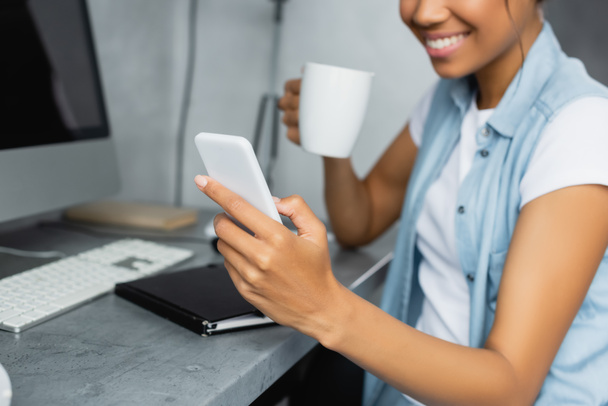 cropped view of african american freelancer holding cup of tea and chatting on smartphone, blurred background - Фото, зображення