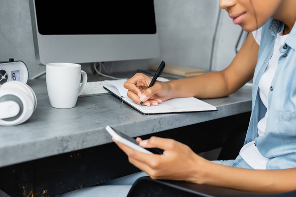 vista ritagliata di freelance afroamericano utilizzando il telefono cellulare e la scrittura in notebook vicino a tazza di tè e computer, primo piano sfocato - Foto, immagini