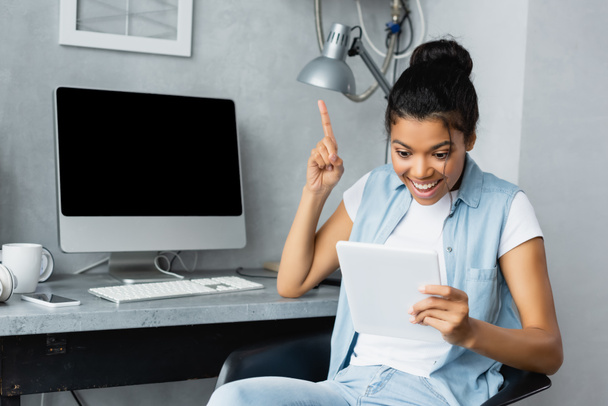 excited african american freelancer showing idea gesture while holding digital tablet near monitor with blank screen - Photo, Image