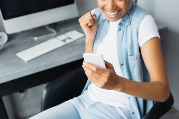 cropped view of african american freelancer holding smartphone and showing win gesture, blurred background - Фото, изображение
