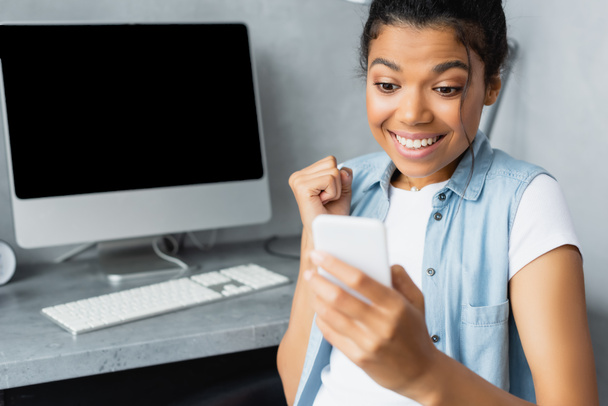 excited african american freelancer showing win gesture near computer monitor and keyboard on blurred background - Foto, imagen