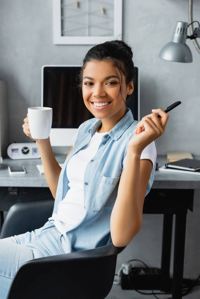 Mutlu Afro-Amerikan serbest çalışanı evde bilgisayarın yanında otururken elinde bir fincan çay ve kalem tutuyor. - Fotoğraf, Görsel