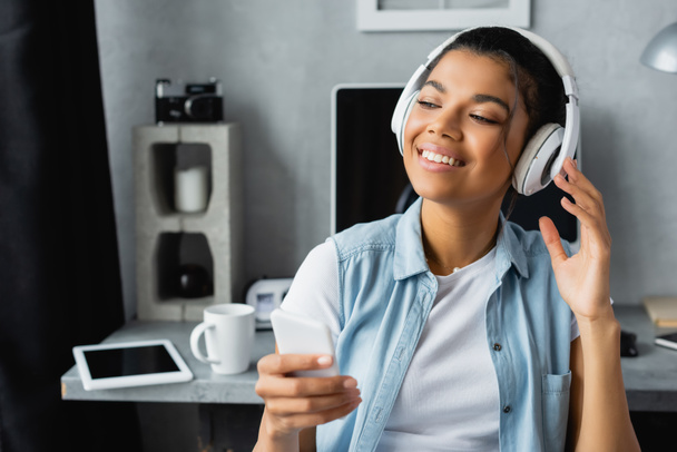 smiling african american freelancer listening music in wireless headphones while holding smartphone at home - 写真・画像