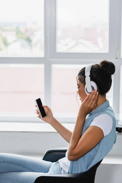 young african american woman in wireless headphones holding smartphone while sitting near window at home - Photo, Image