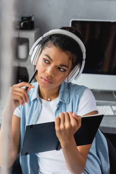 serious, thoughtful african american freelancer in wireless headphones holding notebook and pen, blurred foreground - Фото, изображение