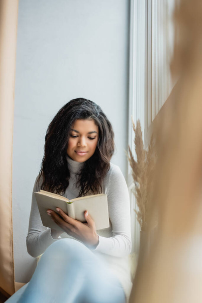young african american woman reading book near window at home, blurred foreground - Photo, image