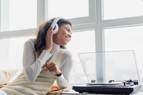happy african american woman in wireless headphones looking through window near record player - Photo, Image