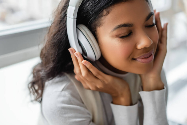 dreamy african american woman listening music in wireless headphones with closed eyes, blurred background - Фото, зображення