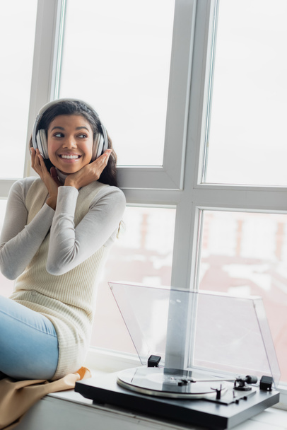 cheerful african american woman touching wireless headphones while listening music near record player on windowsill - Photo, Image