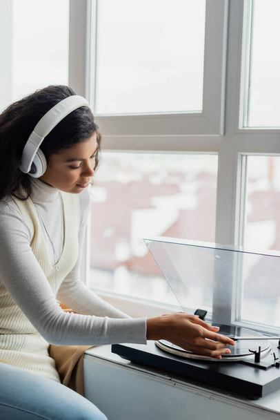 young african american woman in wireless headphones turning on record player near window - Photo, Image