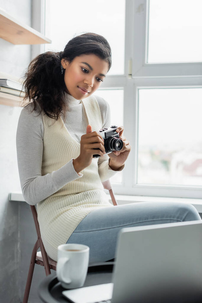 young african american woman holding vintage photo camera while looking at laptop on blurred foreground - Foto, immagini