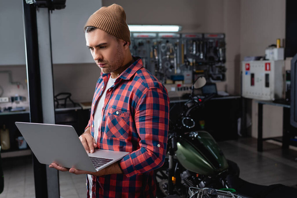 young mechanic in plaid shirt and beanie using laptop in workshop near motorcycle on blurred background - Фото, зображення