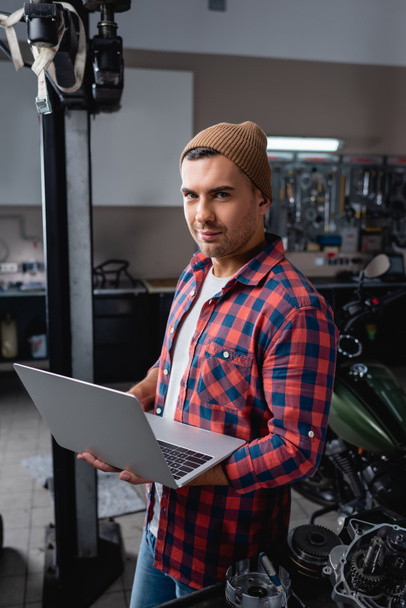 young technician in checkered shirt and beanie smiling at camera while standing with laptop in garage - Фото, изображение