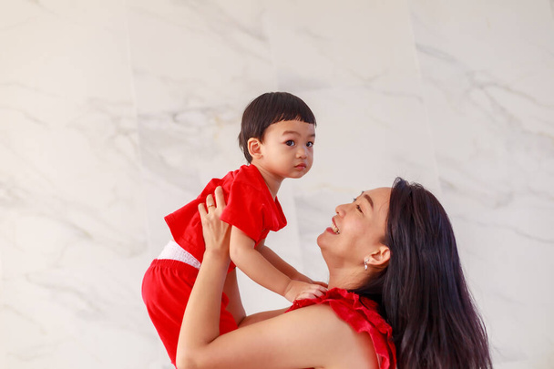 Asyalı bir anne ve küçük oğlu (1 yaşında) beyaz arka planda kırmızı giysiler içinde birlikte oynuyor, gülüyor ve rahatlıyor. Tam boy, kapalı mekan portresi. Mutlu ve eğlenceli oda fikirleri, aile zamanı.. - Fotoğraf, Görsel