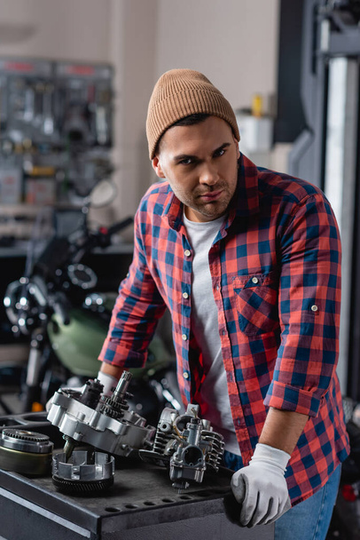 joven mecánico en camisa a cuadros y gorro mirando a la cámara cerca desmontado moto caja de cambios - Foto, Imagen