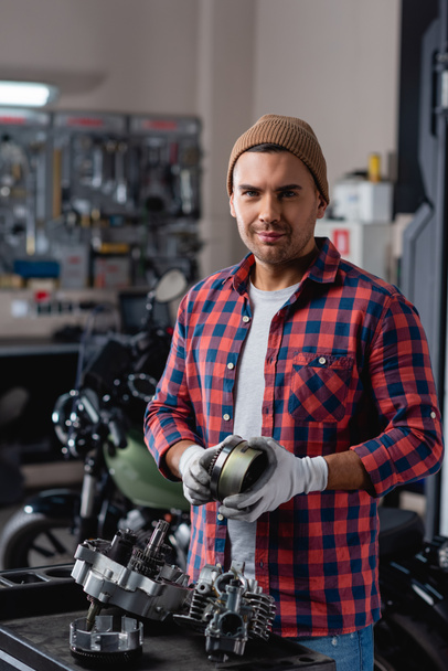 Junger Mechaniker in kariertem Hemd und Mütze blickt in die Kamera neben zerlegtem Motorradgetriebe - Foto, Bild