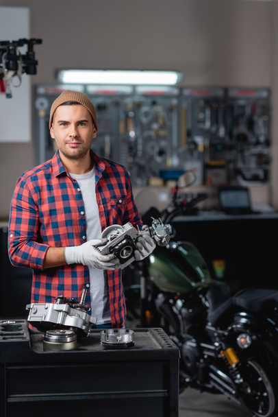 young mechanic in plaid shirt and beanie looking at camera while holding part of gearbox near motorbike on blurred background - Photo, Image