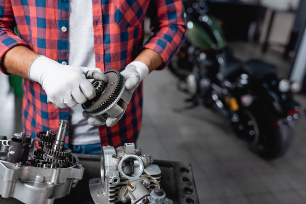 cropped view of mechanic in plaid shirt and gloves holding gearwheel near disassembled motorbike transmission - Foto, immagini