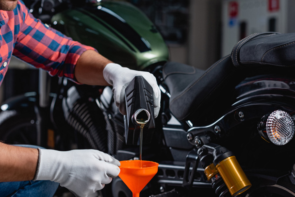 Ausgeschnittene Ansicht eines Mechanikers, der auf verschwommenem Hintergrund Motoröl in einen Trichter neben dem Motorrad gießt - Foto, Bild