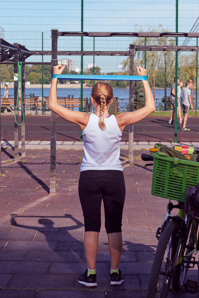 Mujer caucásica hacer ejercicios con banda elástica azul en el patio. Día soleado en el gimnasio exterior. Vista trasera. Foto vertical. - Foto, imagen