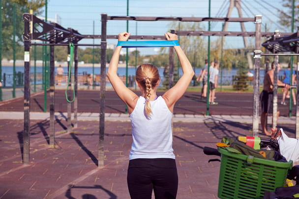 Белая женщина делает упражнения с голубой эластичной лентой на детской площадке. Солнечный день в открытом спортзале. Вид сзади. - Фото, изображение