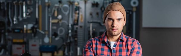young repairman in checkered shirt and beanie looking at camera in garage, banner - Photo, Image
