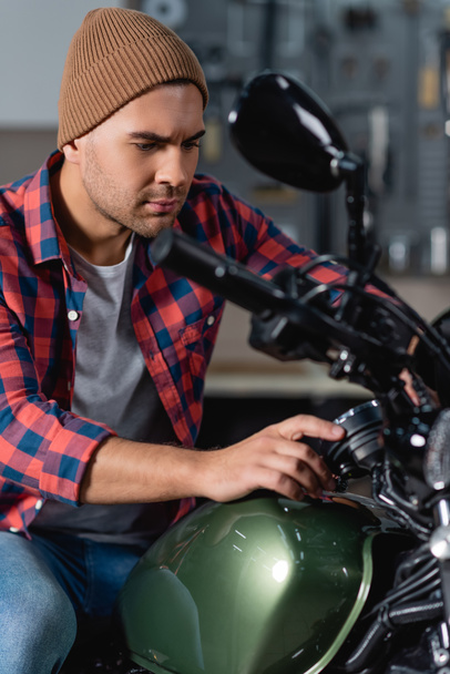 young mechanic in plaid shirt and beanie making diagnostic of motorbike in workshop - Фото, зображення