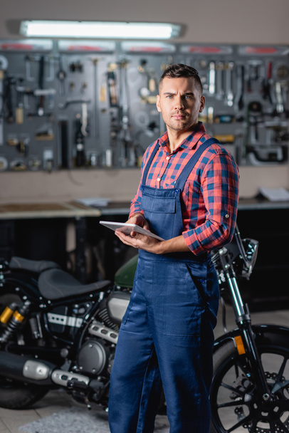 young technician in overalls looking at camera while standing near motorcycle with digital tablet, blurred background - Fotó, kép