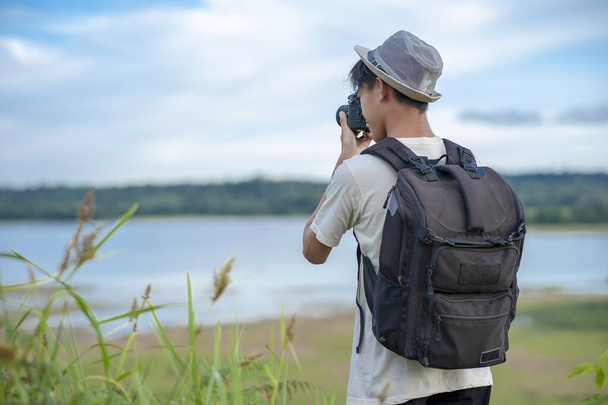 Νεαρός Ασιάτης ταξιδιώτης φορώντας πλεκτό καπέλο με σακίδιο τραβώντας μια φωτογραφία και κοιτάζοντας καταπληκτική λίμνη. Ταξιδιωτική έννοια χαλάρωσης διακοπών - Φωτογραφία, εικόνα