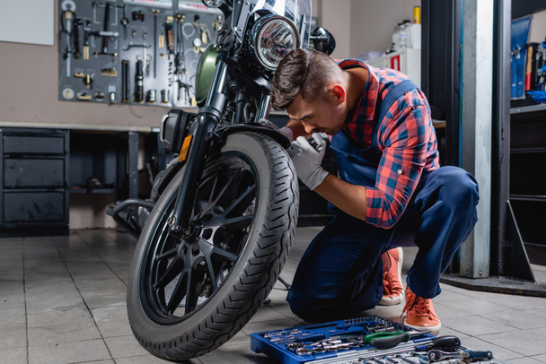joven técnico en overoles haciendo diagnósticos de motocicleta en garaje cerca de caja de herramientas - Foto, Imagen