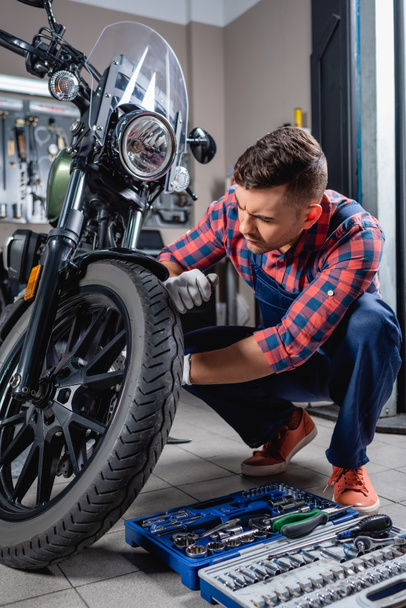 young mechanic in overalls making diagnostics of motorcycle near toolbox in workshop - 写真・画像
