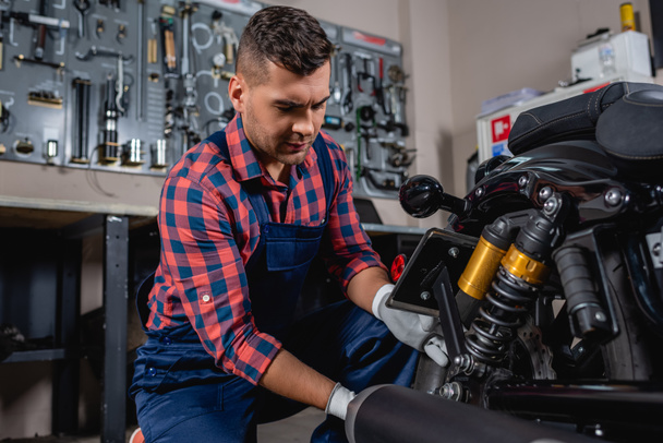 joven mecánico en overol y camisa a cuadros comprobando amortiguador de motocicleta en taller - Foto, Imagen