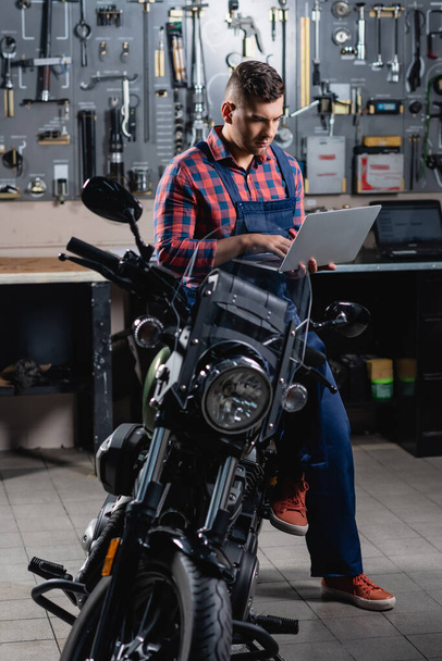 mecánico en overoles usando portátil mientras está sentado en la motocicleta en el garaje - Foto, imagen