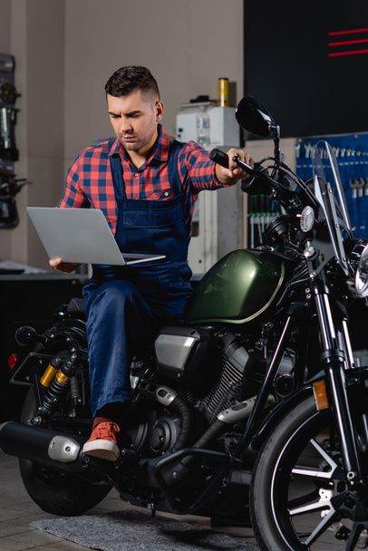 молодой механик в комбинезоне с помощью ноутбука, сидя на мотоцикле в мастерской - Фото, изображение