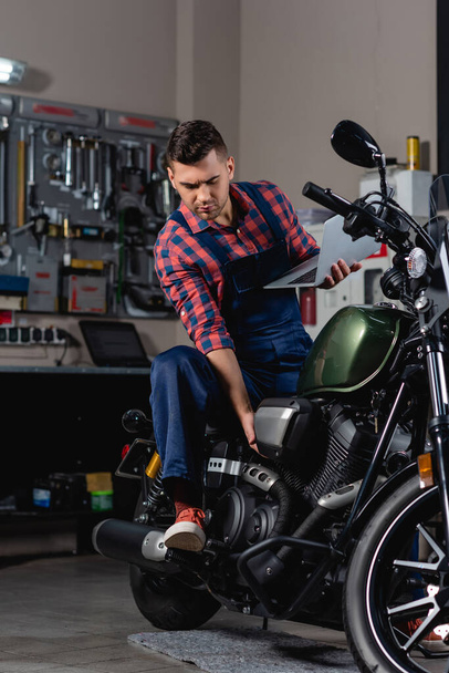 ремонтник в комбинезоне проверяет мотоцикл, держа ноутбук  - Фото, изображение