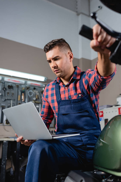 young technician in overalls using laptop while sitting on motorbike in garage - Photo, Image