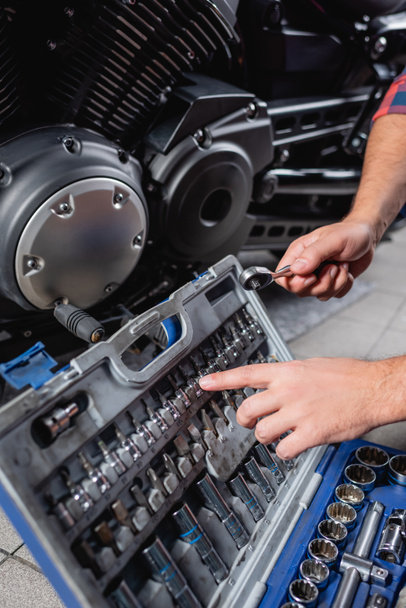 Ausgeschnittene Ansicht des Mechanikers, der mit dem Finger auf Bits für Steckschlüssel im Werkzeugkasten in der Nähe des Motorradgetriebes zeigt - Foto, Bild