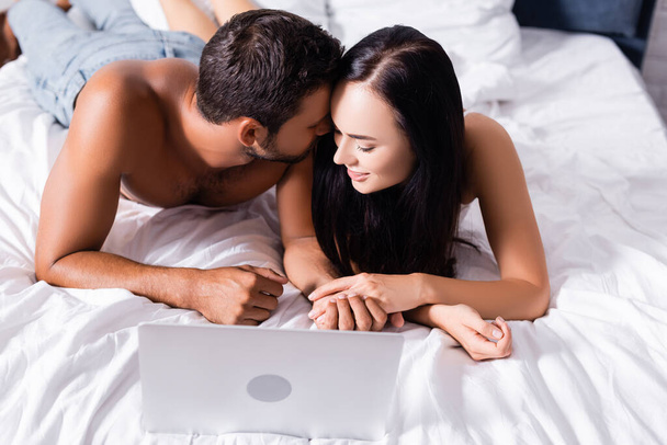 smiling sexy couple holding hands near laptop on bed on blurred background - 写真・画像