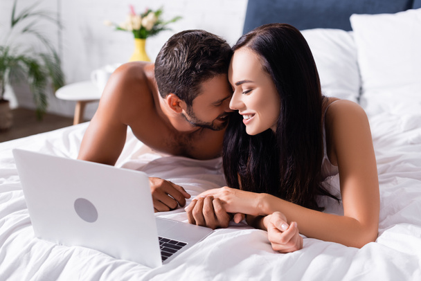 gelukkig paar lachen terwijl het houden van handen in de buurt laptop op wazig achtergrond in slaapkamer - Foto, afbeelding
