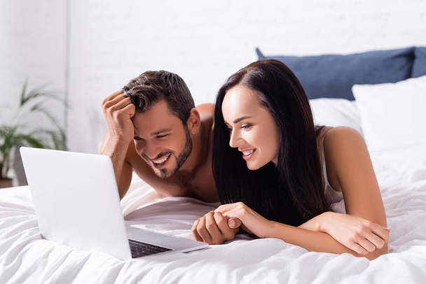 gelukkig jong paar lachen terwijl liggend op bed in de buurt laptop op wazig achtergrond - Foto, afbeelding