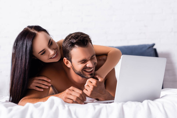happy brunette woman hugging shirtless man laughing near laptop on bed  - Foto, Imagem