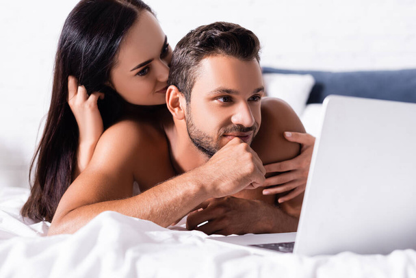 smiling brunette woman hugging man looking at laptop in bedroom on blurred background - Photo, Image