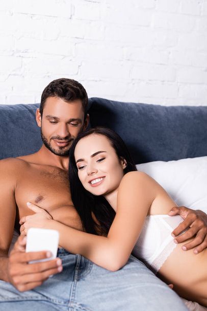 smiling young couple looking at smartphone while lying on bed - Fotografie, Obrázek