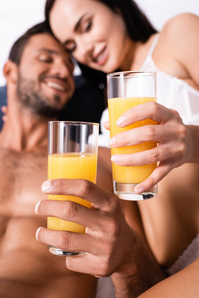 close up view of glasses with juice in hands of happy sexy couple in bedroom on blurred background - Fotografie, Obrázek