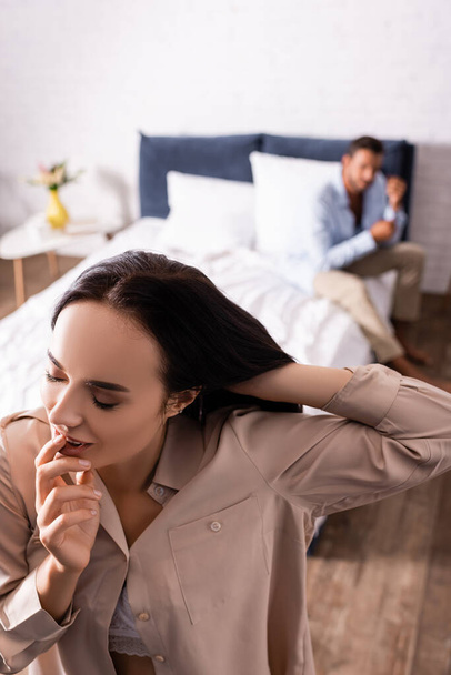 high angle view of sensual woman with closed eyes fixing hair in bedroom with blurred boyfriend on background - Photo, Image