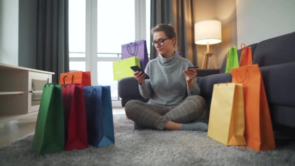 Mujer feliz con gafas está sentado en el suelo y hace una compra en línea con una tarjeta de crédito y un teléfono inteligente. Bolsas de compras alrededor. Compras en línea, tecnología de estilo de vida - Metraje, vídeo
