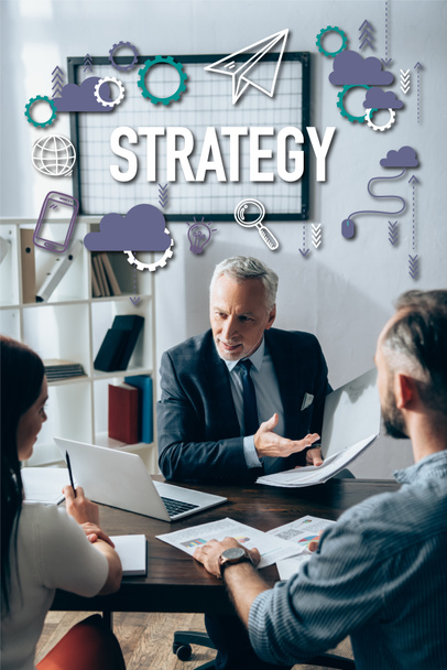 Smiling investor pointing at paper folder near laptop and businesspeople with documents on blurred foreground, strategy lettering  - Фото, зображення