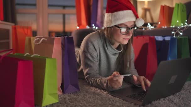 Mujer feliz con gafas con un sombrero de Santa Claus está acostado en la alfombra y hace una compra en línea con una tarjeta de crédito y un ordenador portátil. Bolsas de compras alrededor. - Imágenes, Vídeo
