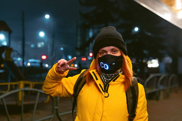 Una chica vestida con una chaqueta de invierno amarilla está parada en una calle de la ciudad por la noche. Hay una máscara en la cara. Máscara con bombillas incorporadas. Iluminación de hielo. para ser diferente. Destacan entre otros - Foto, Imagen